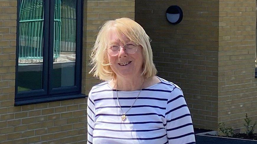 Julie Young, who has short blonde hair, wears a black and white-striped top in front of a cream brick building. She is smiling and wearing glasses.