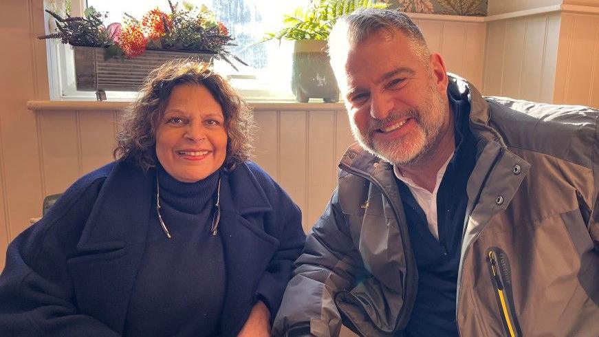 Caroline Gates with brown short curly hair, wearing a royal blue jumper and a royal blue winter coat. She is sat next to Andy Collins, who has short hair that is white and grey, and he is wearing a grey raincoat.