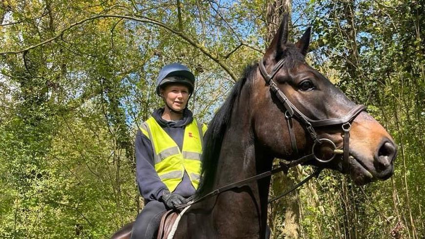 A woman wearing a helmet and a high-visibility jacket riding a brown horse. There are green trees in the background. 