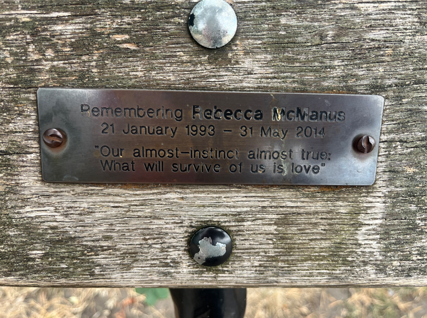 A close-up of a memorial plaque on a bench, which reads: "Remembering Rebecca McManus/ 21 January 1993 - 31 May 2014/ 'Our almost-instinct almost true: What will survive of us is love'".