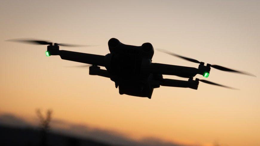 Silhouette of a small drone featuring four small propellers, set against a darkening sky. Two green lights are visible on the drone.