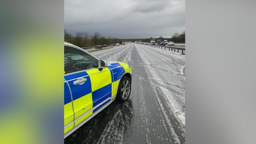 A police car is parked at an angle in the middle of a motorway, blocking it, which has hailstones covering much of the tarmac. Traffic can be seen joining the motorway further ahead and coming in the other direction on the opposite carriageway. 