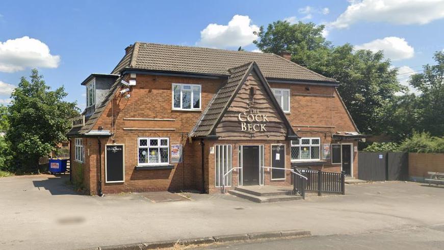 The Cock Beck pub in Leeds, a brick two-storey building with a gable front with two steps leading up to it. It is surrounded by an empty car park. 