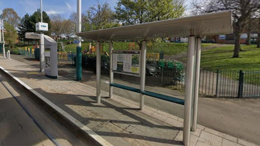 A typical tram stop by a children's park fronted by a racing green- coloured gate, railings and bushes. The tram stop has a pale grey, grooved standing area raised by a high kerb. The stop itself is grey shiny steel, with a full glass back and a green-coloured bench on a slight tilt. 