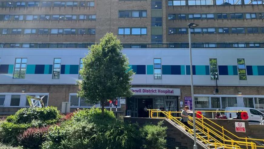 Outside of Yeovil district hospital's main entrance, with yellow steps and green bushes. The building is brown bricks