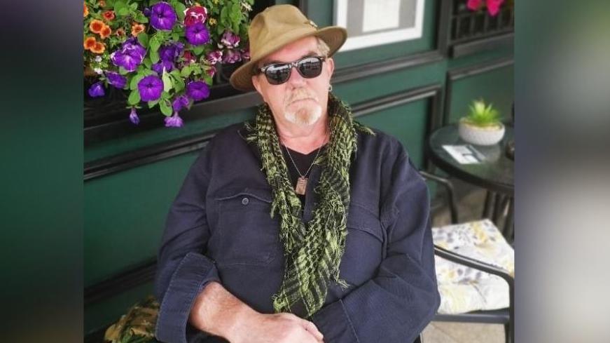 A man sat outside a bar or restaurant. He is wearing a navy shirt, a green chequered scarf, dark sunglasses and a brimmed hat. He is beside some colourful flowers in a window box.   