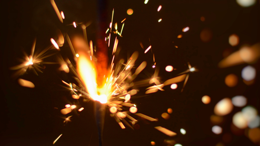 A close-up of a sparkler which is alight and has yellow flames against a black background.