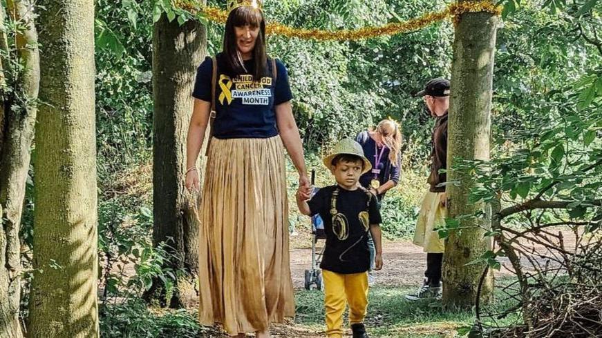 Lisa Radcliffe with shoulder length black hair, wearing a black T-shirt and a long gold pleated skirt, holding the hand of son George, aged three, who is wearing a large yellow hat, a black T-shirt and yellow trousers, while they walk through woodland