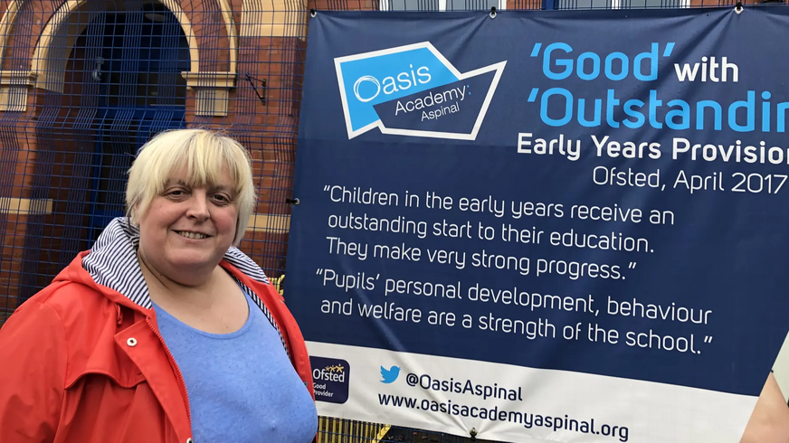 Jackie Pearcey has a red jacket, sky blue sweater and bobbed blonde hair. She is standing outside Gorton Oasis Academy.