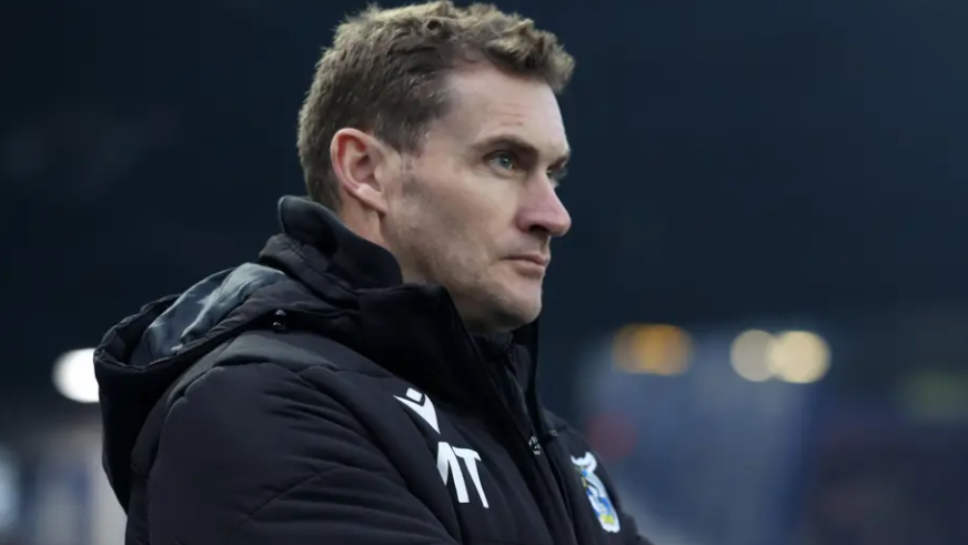 A close-up of Matt Taylor in a thick black winter coat against a blurred background, looking thoughtfully into the distance to the left of camera