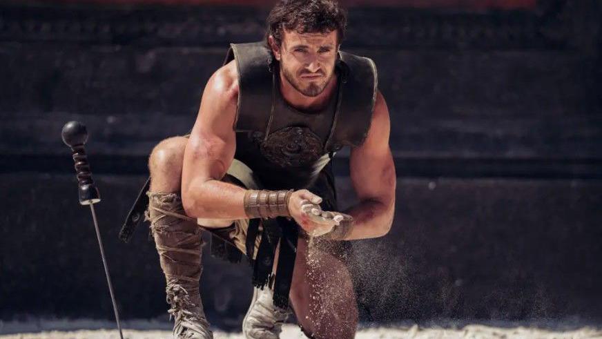 Paul Mescal plays Lucius, second Gladiator film. A man crouches, kneeling on the ground wearing a leather upper body sleeveless armour, calf length boots and rubs his hands together. Beside him, a sword embedded in the ground