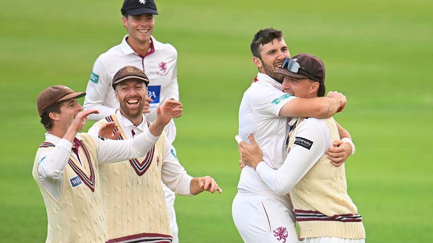 Five cricket players in white cricket kits are laughing and hugging each other on a pitch. The two on the right of the image are embracing, while the other three are all smiling. Two of them have their arms partly raised. 