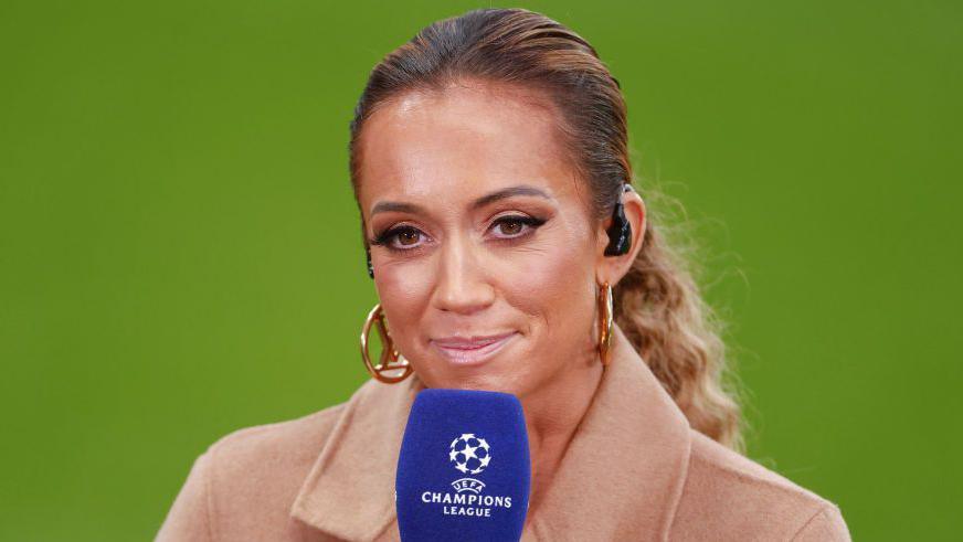 Kate Abdo presenting the Champion's League pitchside. She has blonde hair in a ponytail and is wearing a camel coat and large gold hooped earrings