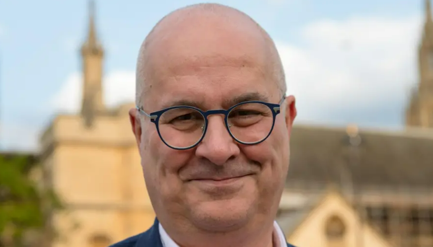 Image shows Iain Dale. He is bald and wearing black, round-rimmed glasses, and a blue suit. He is standing in front of the Palace of Westminster, which is blurred in the background.