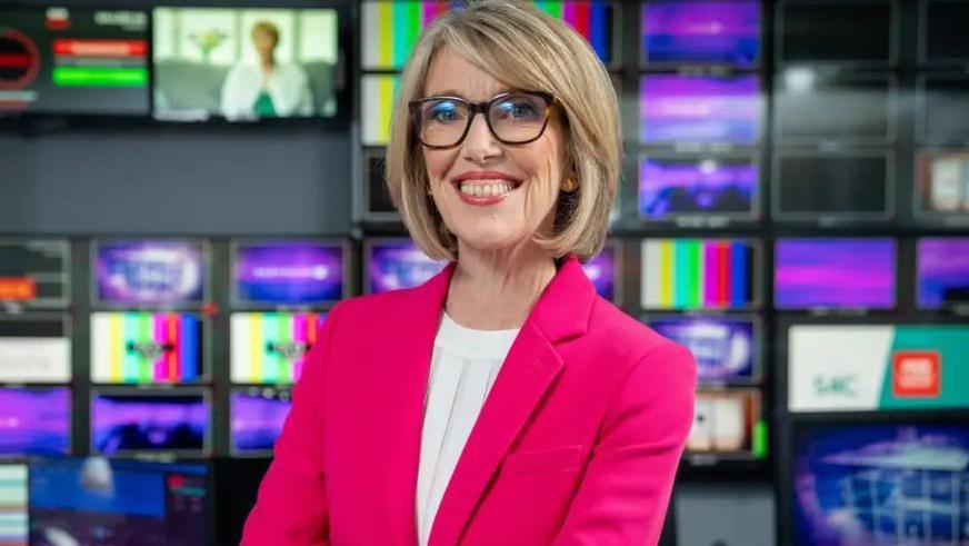 Bethan Rhys Roberts wearing a pink jacket and glasses. She is sat in from of several screens in a tv gallery room.