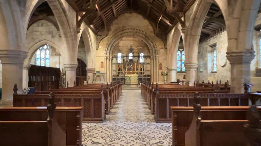 Inside St Edith's church there is a tiled floor consisting of an intricate mosaic design which stretches all around the building. Several lines of wooden pews line the way to the altar which includes brass figures. Light from the stained glass windows seeps through, illuminating the edges of the church. 