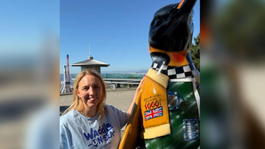 Amy Dearing is standing to the left of the image with her arms resting on a penguin sculpture on the right. She has shoulder length blonde hair and is wearing a blue T-shirt that says Waddle-on-Sea on the front.