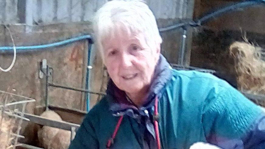 Sue, a woman with short white hair, she wears a green jacket with a navy hood and smiles, she is in a farm building.
