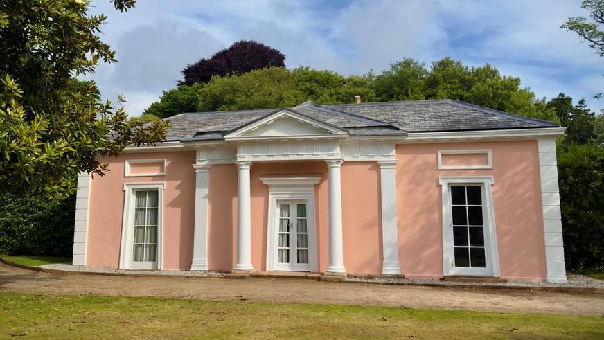 A house painted a salmon pink colour with white accents. The roof is grey.
