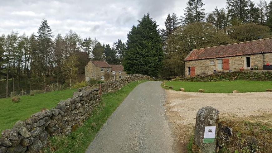 A lane in Urra following a dry stone wall with stone houses and outbuildings on either side.