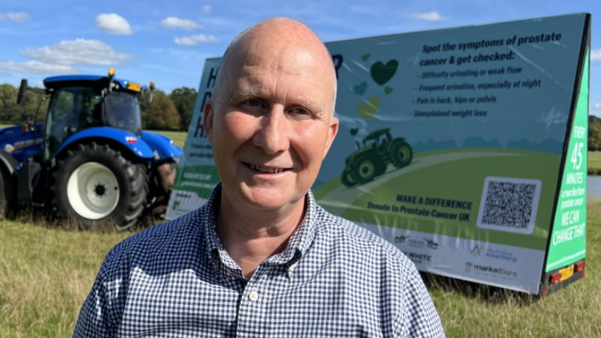 Andrew Gibson standing in front of a blue tractor with a trailer that is advertising prostate cancer figures, the symptoms and urging people to get checked.