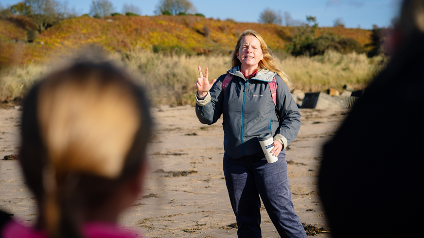 Bridie Melkerts is talking to a group of children on a beach with the image taken from behind the heads of two of the children. She is wearing jeans and a blue top and has long blonde hair. 