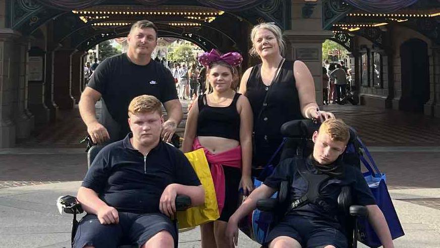 Bradley, Laura and Boe all standing behind Ralph and Rudi, who are sitting in their wheelchairs, at a Disney resort. They are smiling for this family photo, taken on a warm day.