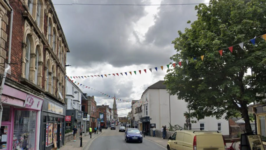 Fisherton Street with shops, cars, pedestrians, a tree in the foreground on the right and a church spire in the background