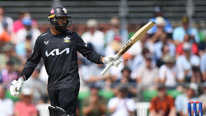 Ryan Patel raises his bat to the crowd after scoring a half-century. He is wearing an all black Surrey One-Day Cup kit
