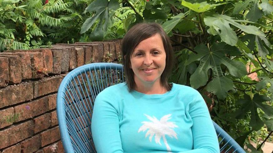 Yn y llun yma mae'r Athro Eleri Rosier yn gwenu. Mae'n gwisgo siwmper glas. Mae ganddi wallt byr brown. Mae hi'n eistedd ar gadair yn yr ardd.