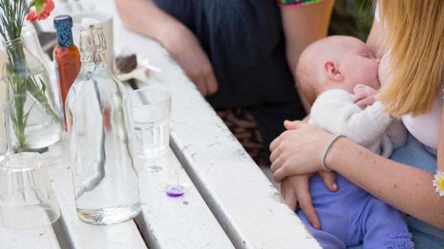 The main part of the image is a white wooden table with glasses and a bottle of water on it, also a vase with flowers. Sitting at the bench is a woman with reddish blonde hair although her face is obscured. Her baby is feeding from her right breast - the child is about 4-6 months old and wear a white top and purple leggings.