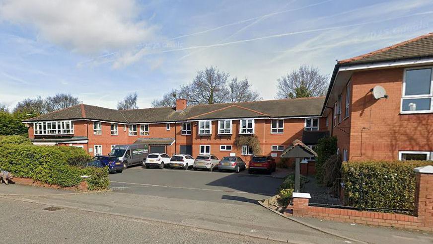 The exterior of the Aaron Court Care Home on a clear day with hedges at the side of the entrance to the car park and building.