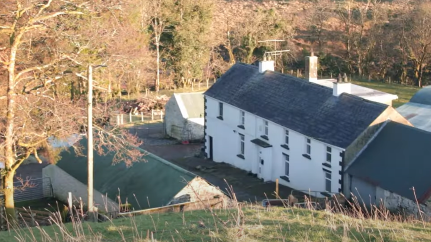 The white farmhouse which has been in the McGoldrick family for generations is pictured from an aerial view. It is white and rectangular shape with black windowsills, cornerstones and a black roof. There is a white brick shed with a green roof, and there is another white shed. The house is surrounded by trees and green fields.