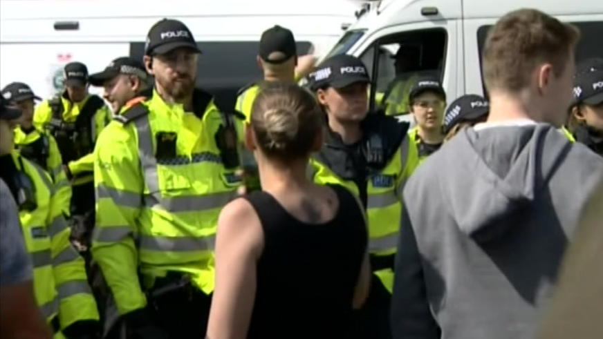 A woman in a black vest top faces police with a man in a grey hoodie stood to her right. A line of uniformed police officers stand in front of her with a van in the background.