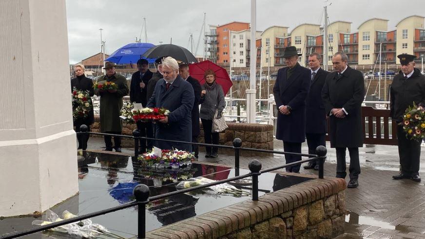 People dressed in dark clothing in including winter coats and suits laying a wreath on a memorial. It is a rainy day.