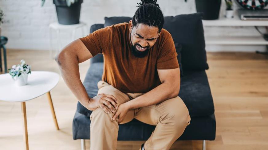 Man on grey sofa chair, wearing brown t-shirt and trousers, clutching right knee in pain from osteoarthritis