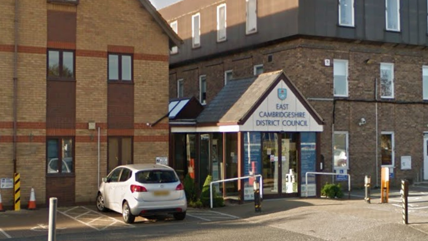 2 buildings are in the picture. One is on the left, and one is on the right. In the middle is a small building which is an entrance to the council building. It has a sign above which says 'East Cambridgeshire District Council'. 