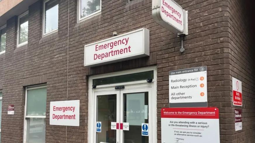 A picture of the front doors of the Emergency Department in St Helier, Jersey. The hospital building is made up of brown bricks, and the doors are glass. 