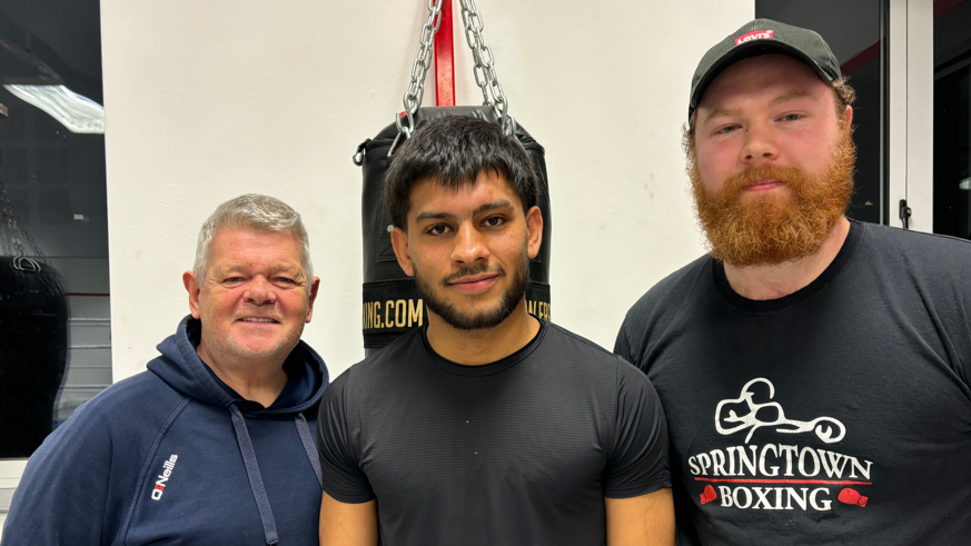 Shows Mujeeb in the centre in front of a punch bag with a man in a hooded top on the left and another with a Levis baseball cap, red beard and black t-shirt. 