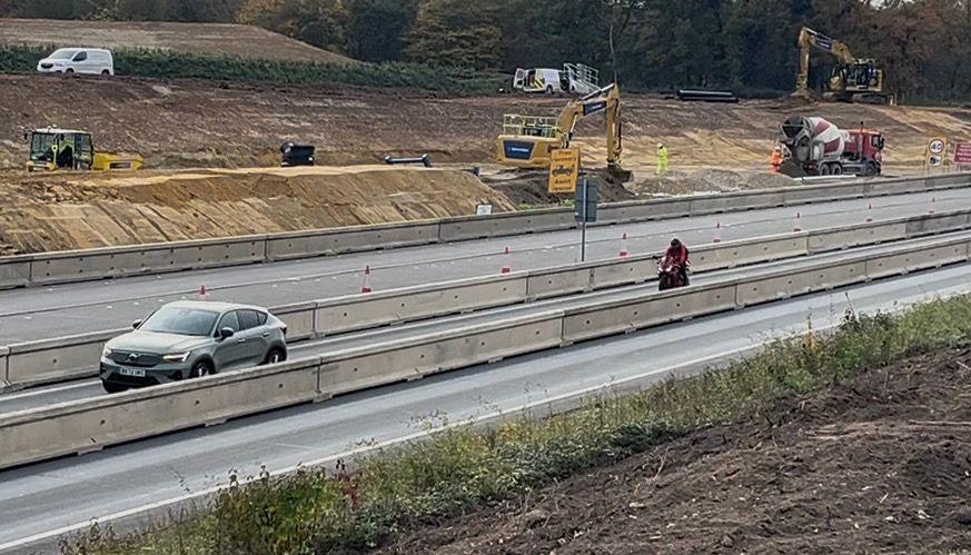 A car and a motorbike travelling right to left on a dual carriageway, with temporary hoardings creating one narrow lane. Behind them, a bank of sand is piled in parallel with the road, with a digger and mixer lorry parked there.