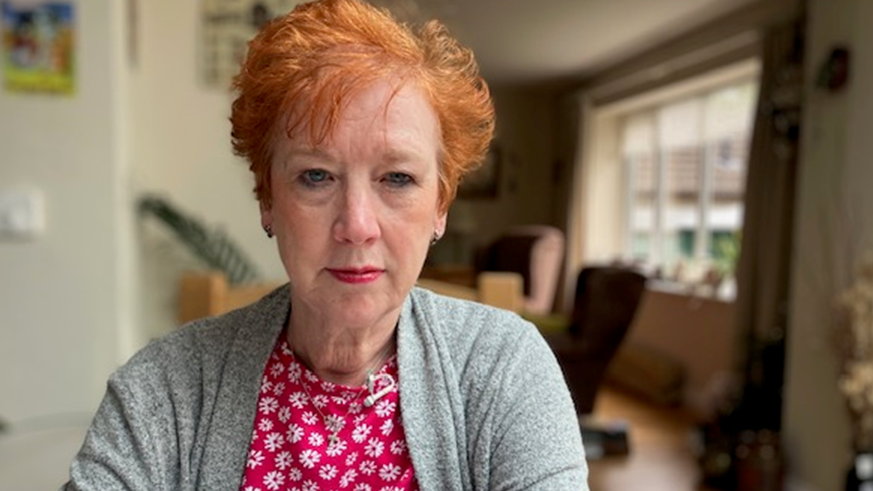 Anita Tonks, wearing a red top with a grey cardigan, sat at a table in her home looking directly into the camera.