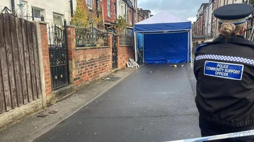 A forensic tent stands in a back alley. A police community support officer stands with her back to the camera in the foreground.