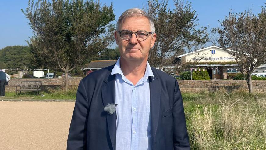 A man with grey hair wearing a dark blue blazer and light blue shirt and round black-rimmed glasses looks at the camera while stood next to a petanque court and in front of a community building.