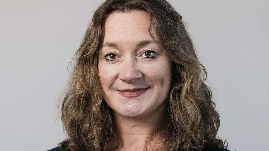 Daphne Caine, who has long, wavy, light brown hair, against a grey background.
