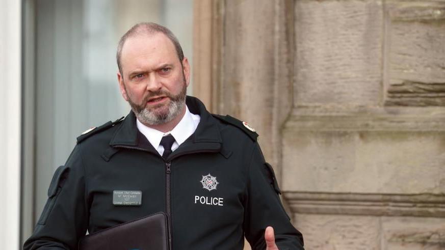 Mark McEwan talking beside a building wall, carrying a black folder. He is wearing a shirt and tie under a black police jacket.