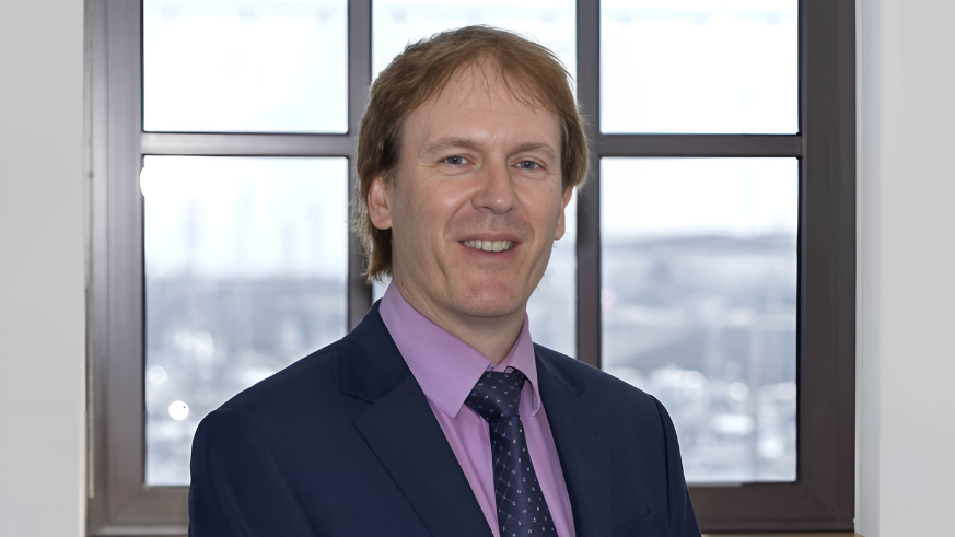 Richard Hemans wearing a dark blue suit and tie and a purple top. He is stood in front of a window.