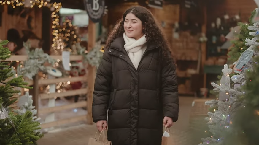 A woman in a black coat with long dark curly hair holding two paper shopping bags in a Christmas-themed setting with fairy lights and Christmas trees.