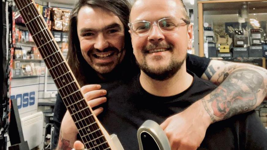 Sam Orr is smiling as he stands behind Mark Wood in Sam's guitar shop. Mark Wood is also smiling as he holds his guitar, with the neck of the guitar and the frets visible.