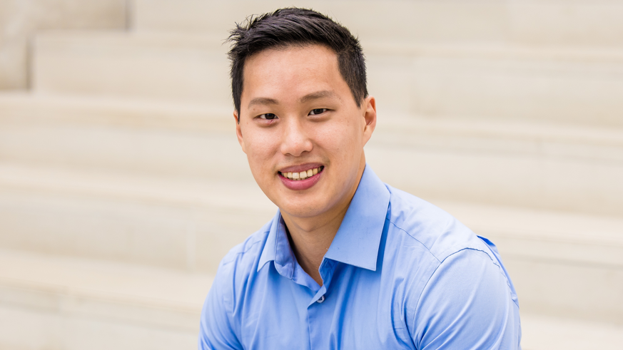 A headshot of a man smiling at the camera. He has black hair and a light blue shirt on.