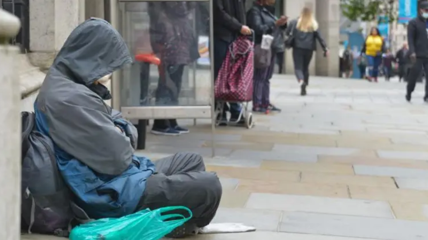 A rough sleeper sitting by a bus stop on a busy town pavement, wearing a rain coat with the hood up and leaning against a rucksack with a plastic bag beside him